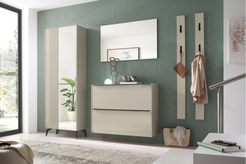 Kaschmirfarbene Flurmöbel mit Glasfront und schwarzen Details. Abgebildet sind Kommode, Spiegel, Garderobenpaneel und Schrank vor einer grünen Wand.