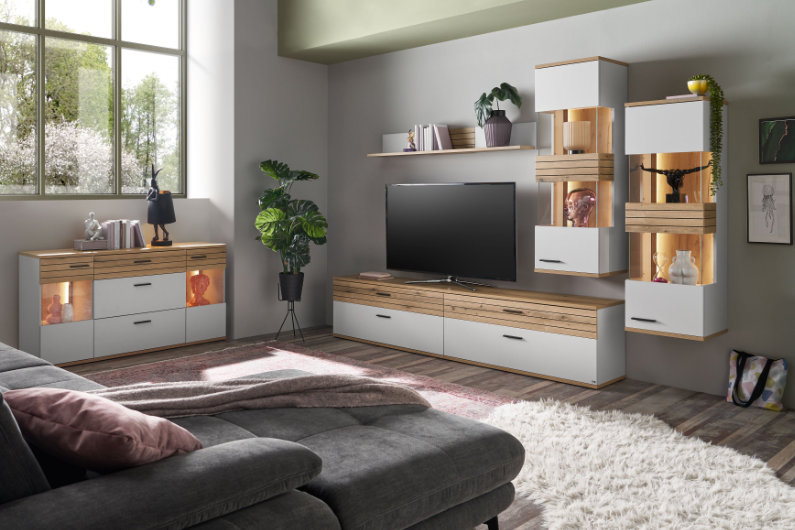 Wohnzimmer mit einer beleuchteten Wohnwand und einem Sideboard in Lichtgrau. Mit Absetzungen in Eiche
